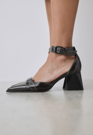 Zapato negro de cuero con puntera puntiaguda y tacón grueso, con correa al tobillo y hebilla, usado en pie desnudo sobre un fondo liso.