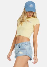 T-shirt jaune court avec un col rond et des manches courtes, associé à un short en denim bleu clair avec des déchirures et un ourlet roulé. Casquette bleue.