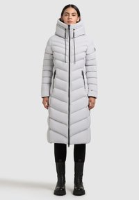 Hosszú fehér pufferek coat magas gallérral, chevron steppelésű, elülső cipzárral és oldalsó zsebekkel. Fekete magas csizmákkal és leggings-szel párosítva.