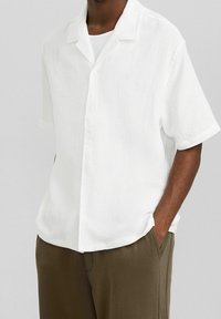 Camicia bianca a maniche corte con tessuto testurizzato, apertura con bottoni e colletto classico, abbinata a pantaloni marroni.