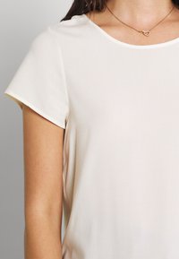 Lys beige kortermet topp med rund hals. Glatt tekstur, uformell design, med en subtil glans. Kombinert med et delikat hjertehalssmykke.