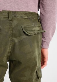Spodnie typu cargo w kolorze oliwkowej zieleni, wyposażone w boczne kieszenie, płaski pas oraz trwały materiał. Wzór zawiera delikatny kamuflaż.