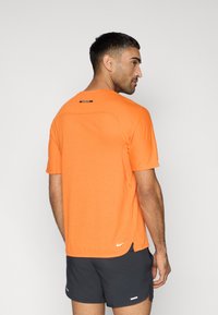 Λαμπερό πορτοκαλί αθλητικό t-shirt με κοντά μανίκια, υφασμένη πίσω πλάτη και μικρό λευκό λογότυπο στο κάτω αριστερό μέρος. Φορεμένο με σκούρα σορτς.