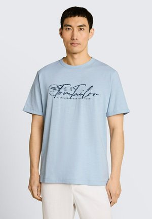 Man draagt een lichtblauw T-shirt met korte mouwen met de tekst "Tom Tailor" en een witte broek, staand tegen een effen lichte achtergrond.