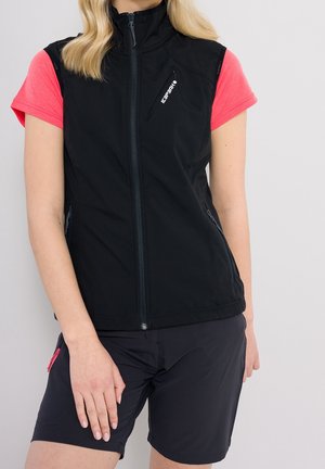 Femme portant un gilet noir sans manches zippé sur un t-shirt rose à manches courtes et un short noir, debout la main gauche posée sur sa cuisse.