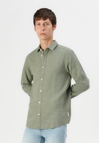 Camisa de manga larga abotonada en una mezcla de lino verde oliva, con cuello clásico, botones blancos y dobladillo curvado. Ajustada y con una textura ligera.