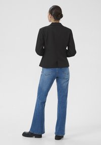 Schwarze maßgeschneiderte Blazerjacke mit figurbetontem Design, kombiniert mit hellblauen Schlagjeans. Das Outfit zeichnet sich durch einen Rückenschlitz und glatte Texturen aus.