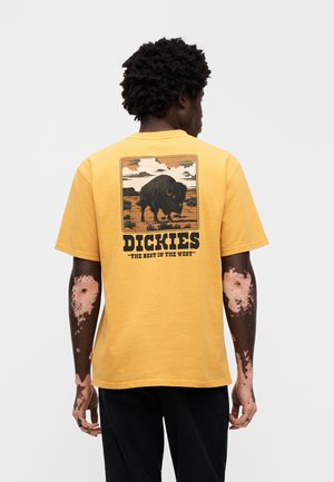 Gul bomulls-t-skjorte med en stor grafikk av en bison, omkranset av landskapsbilder, med teksten "Dickies" og en slagord nederst.