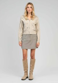 Haut à capuche zippé beige avec des embellissements floraux, associé à une jupe grise et des bottes beiges montantes. Design épuré, texture douce.
