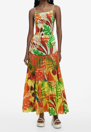 Kobieta w bezrękawnej, warstwowej sukni maxi z wielokolorowym tropikalnym motywem liści oraz białych sandałach na platformie.