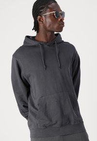 Charcoalgrauer Hoodie aus einem Baumwoll-Mischgewebe, mit einer vorderen Kängurutasche und einer Kapuze mit Kordelzug. Das Model trägt runde schwarze Sonnenbrillen.