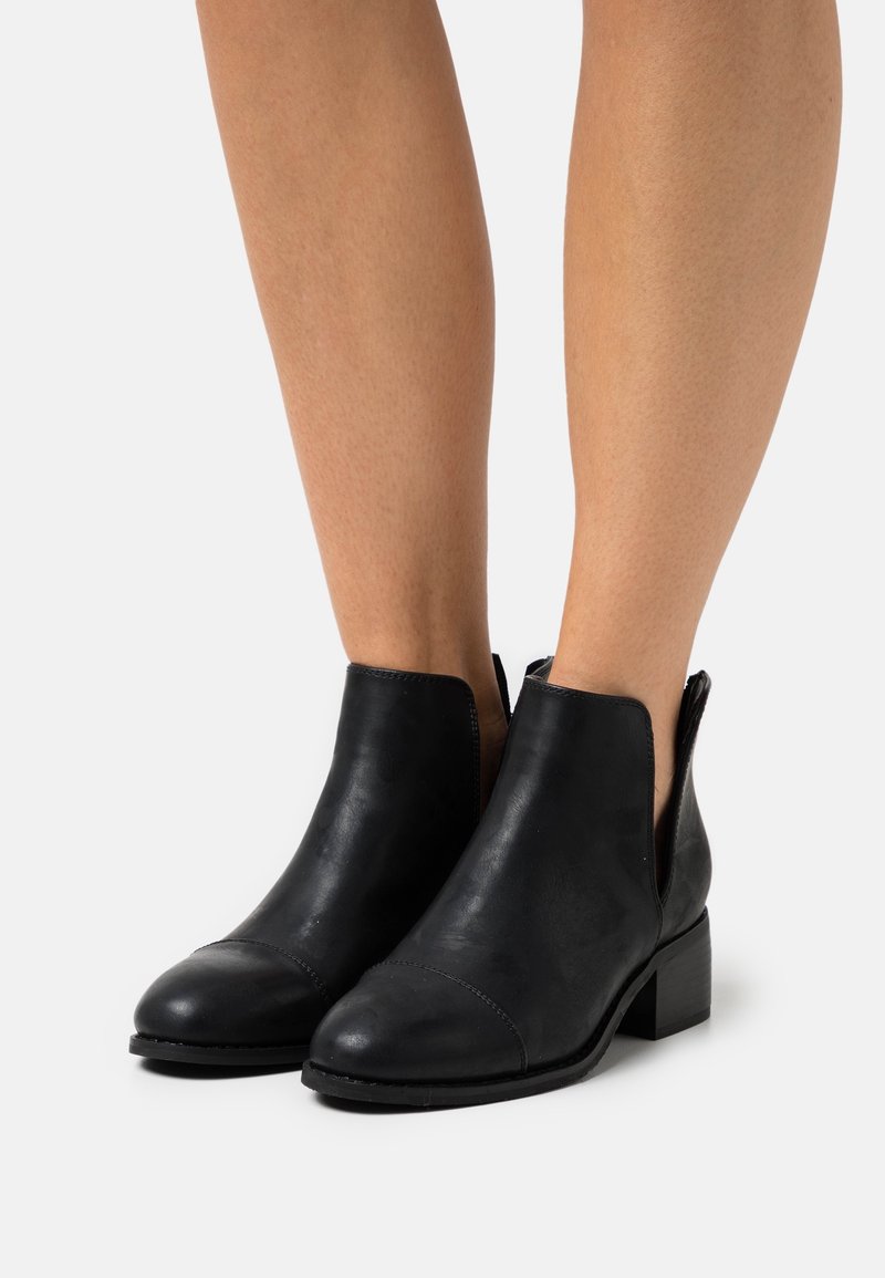 Bottes cheville en cuir noir avec un bout arrondi, un design latéral découpé et un talon carré bas. Texture lisse avec un détail de couture minimal.