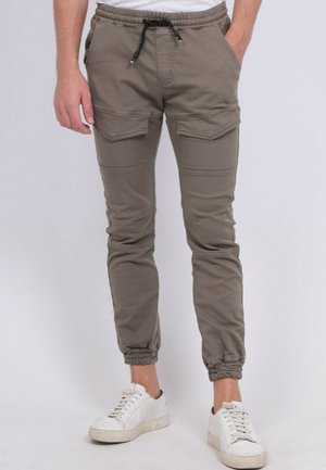 Homme portant un pantalon de jogging cargo gris avec des poignets élastiques et un cordon noir, associé à des baskets blanches et une chemise blanche.