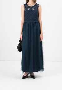 Kvinna i en marinblå ärmlös spetsklänning med genomskinlig kjol, svarta kitten heels, håller en liten svart handväska.