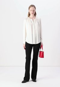 Blusa bianca a maniche lunghe con dettagli plissettati, abbinata a pantaloni neri a palazzo, mentre tiene una piccola borsa rossa. Tessuto morbido, design minimalista.