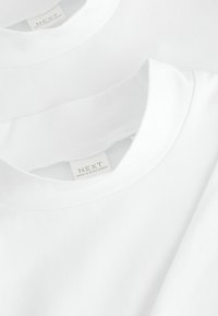 Weiße Baumwoll-T-Shirts gestapelt, mit Rundhalsausschnitten und einer dezenten Textur. Das Etikett zeigt die Marke "NEXT, hergestellt in Bangladesch."