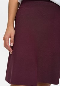 Detailný záber ruky oddychujúcej vedľa sukne s dĺžkou po kolená z tmavo purpurového textúrovaného materiálu, nosenej s bielym topom.