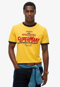 Camiseta de algodón amarilla con ribete negro y texto gráfico rojo "SUPERMAKE". Llevada con una chaqueta de mezclilla azul atada a la cintura. Jeans oscuros visibles.