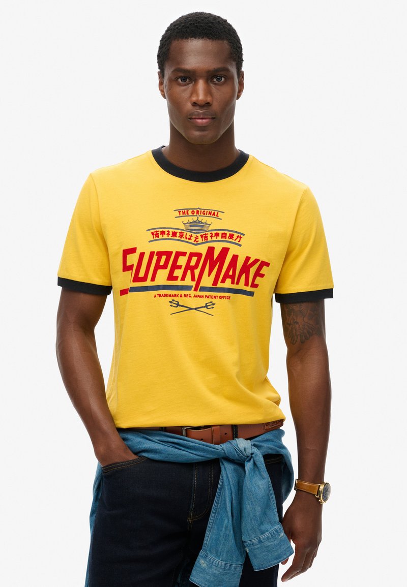 Camiseta de algodón amarilla con ribete negro y texto gráfico rojo "SUPERMAKE". Llevada con una chaqueta de mezclilla azul atada a la cintura. Jeans oscuros visibles.