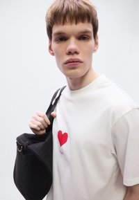 Λευκό βαμβακερό T-shirt με κόκκινο γεωμετρικό γραφικό καρδιάς, σε συνδυασμό με μια μαύρη υφασμάτινη τσάντα ώμου. Απλός, καθαρός σχεδιασμός.