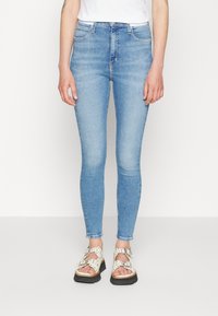 Osoba oblečená v světlých modrých skinny jeans a bílých sandálech se sponou stojí na bílém pozadí.