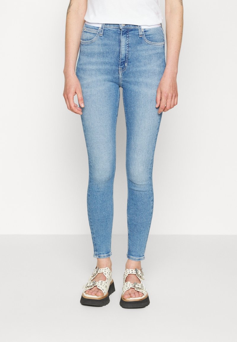 Osoba oblečená v světlých modrých skinny jeans a bílých sandálech se sponou stojí na bílém pozadí.