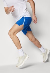 Pantalones cortos deportivos azules con un acento azul más claro, que presentan un dise�ño de lado partido. Llevados con una camiseta blanca y zapatos de colores claros con una superficie texturizada.