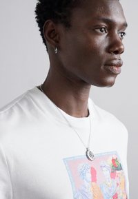 Tricou alb din bumbac cu un print grafic colorat care înfățișează figuri clasice; un pandantiv rotund din argint și cercei tip hoop completează ținuta.