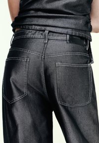 Jean en denim noir avec une texture lisse, doté d'un design classique à cinq poches et de coutures blanches contrastantes.