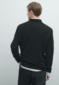 Zwarte wollen trui met een geribde kraag en manchetten, met een ontspannen pasvorm. Gekozen over een lichtgekleurde blouse, met een zachte textuur.