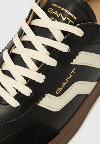 Schwarzer Ledersneaker mit cremefarbenen Schnürsenkeln und seitlichen Akzenten. Verfügt über goldene Markenveredelung und eine strukturierte braune Gummisohle.