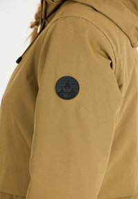 Brun vattentät jacka med texturerad yta, huva och en svart cirkulär patch med ett bergslogo på vänster ärm.