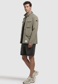 Jachetă ușoară khaki cu patru buzunare frontale, guler înalt și fermoar contrastant. Purtați cu pantaloni scurți închiși la culoare și adidași. Textură netedă.