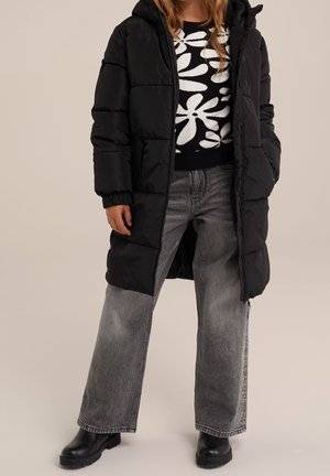 Personne portant un manteau matelassé noir par-dessus un pull à motif floral noir et blanc, un jean gris à jambes larges et des bottes noires.