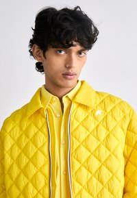 Casaco acolchoado amarelo com colarinho pontiagudo e fecho à frente, combinado com uma camisa amarela abotoada, apresentando um pequeno logotipo no casaco.