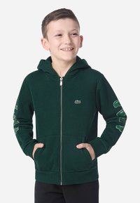 Fleece verde com fecho, apresentando um bolso canguru, punhos canelados e bordado do logotipo no peito esquerdo e na manga. Tecido macio e texturizado.