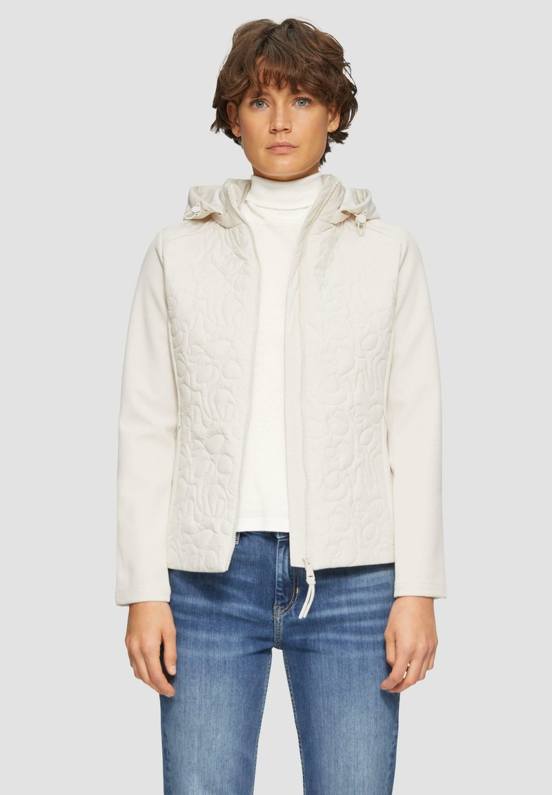 Veste beige clair avec un devant matelassé et des manches douces, dotée d'une capuche et d'un zip frontal. Portée sur un haut blanc et un jean bleu.