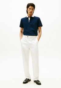 Camisa polo navy con cuello blanco, combinada con pantalones blancos y zapatos negros. La polo tiene un logo en el pecho y mangas cortas.