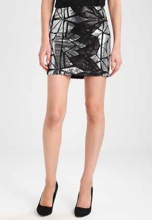 Femme portant une mini-jupe à sequins géométriques noirs et argentés et des talons hauts noirs, debout devant un fond clair uni.