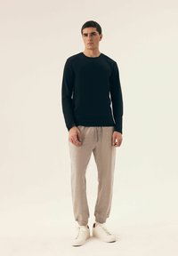 Schwarzes Langarmshirt und hellgraue Jogginghose. Das Shirt hat einen runden Ausschnitt; die Hose hat elastische Bündchen und einen Kordelzugbund. Weiße Sneakers.