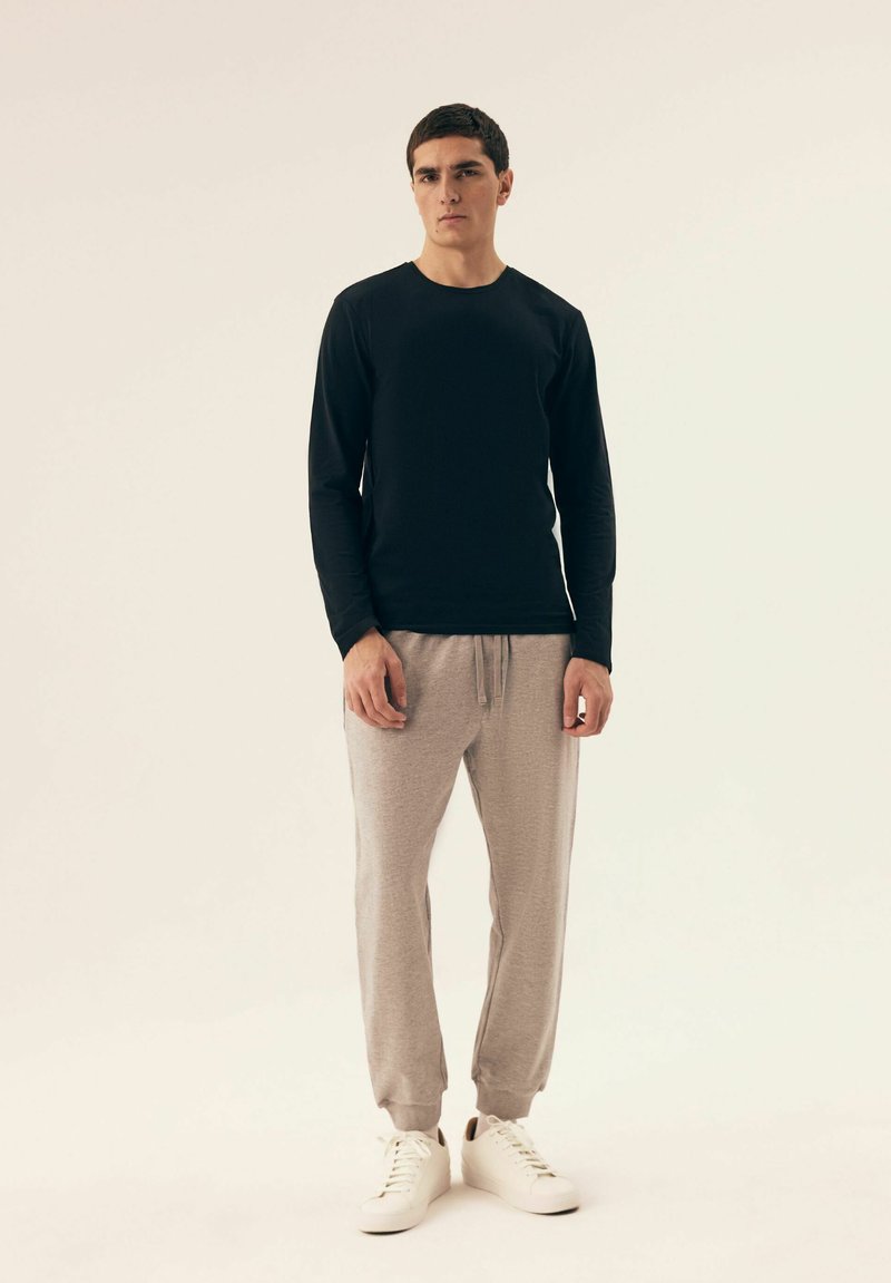 Schwarzes Langarmshirt und hellgraue Jogginghose. Das Shirt hat einen runden Ausschnitt; die Hose hat elastische Bündchen und einen Kordelzugbund. Weiße Sneakers.