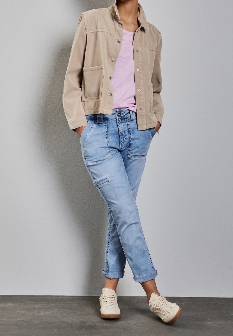 Chaqueta de mezclilla beige con dos bolsillos frontales sobre una camiseta rosa clara; combinada con jeans de lavado claro y puños y zapatos beige con cordones.