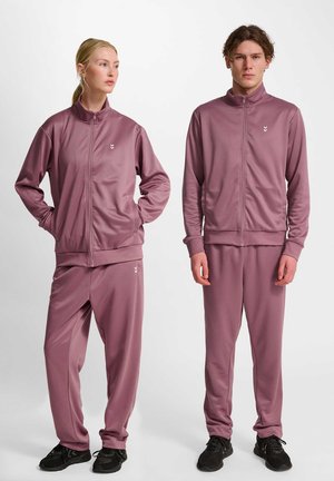 Dwie osoby ubrane w pasujące do siebie mauveowe dresy na suwak i czarne trampki, stojące na jednolitym białym tle.