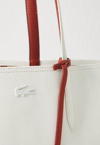 Sac cabas blanc texturé avec anses rouges contrastantes et bordures, avec un petit logo crocodile blanc à l'avant.