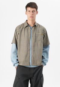 Young man stands confidently, wearing a short-sleeve plaid shirt over a long-sleeve blue top, paired with black pants.