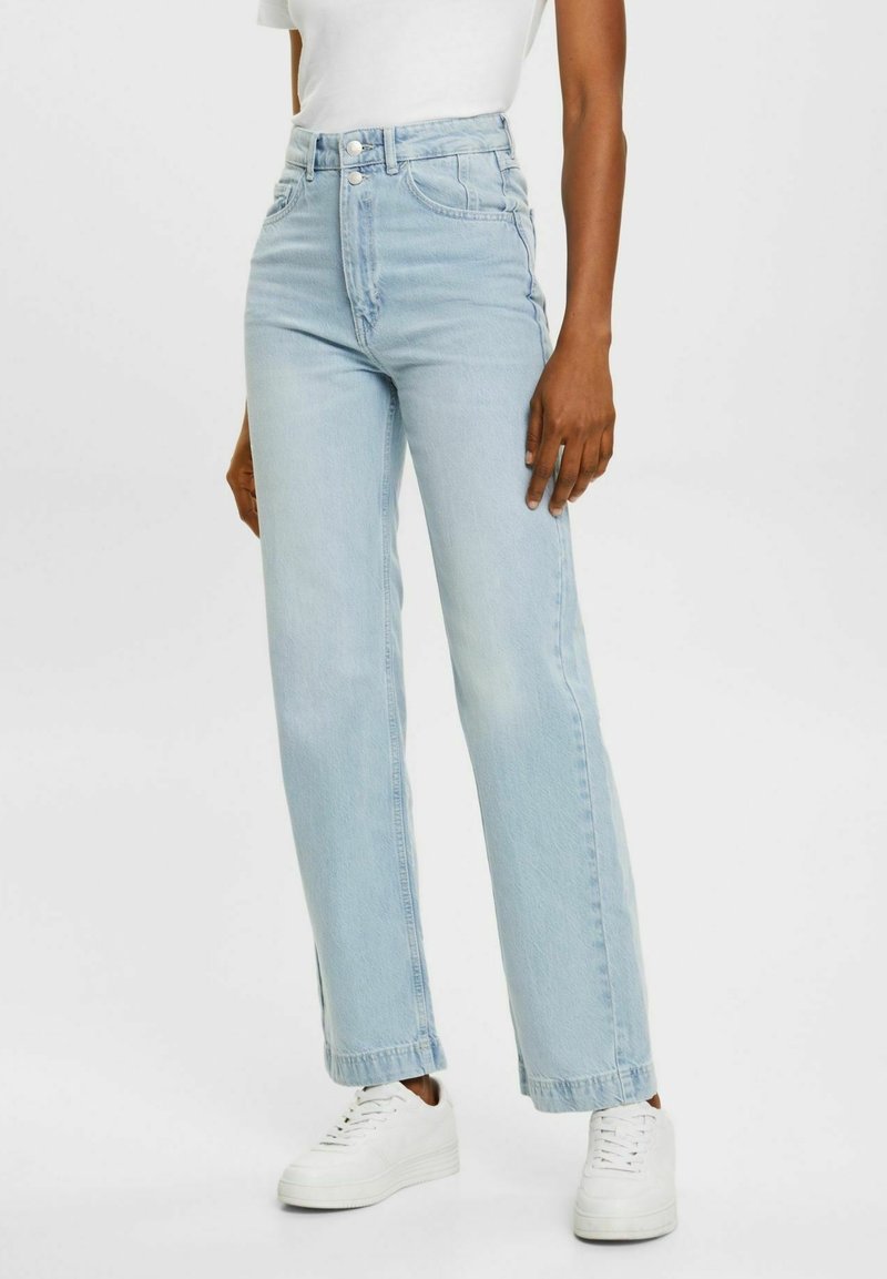 Personne portant un jean bleu clair taille haute droit, des baskets blanches et une chemise blanche à manches courtes rentrée.