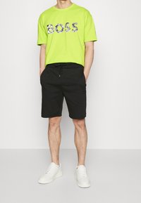 Neon zelené tričko s krátkým rukávem se třpytivým logem "BOSS", spárované s černými šortkami s elastickým páskem a bílými teniskami.