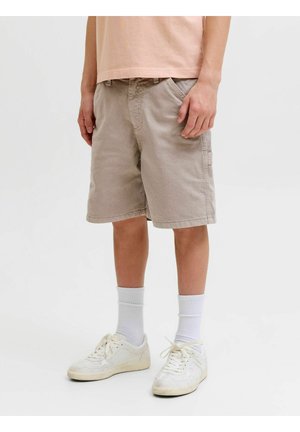 Personne portant un short beige mi-long, des chaussettes blanches montantes et des baskets blanches à lacets, debout sur un fond uni.