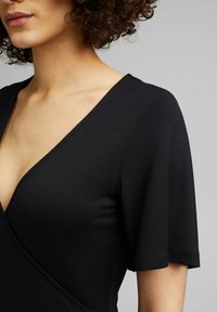 Femme portant un haut noir à manches courtes avec un col en V, aux cheveux bouclés et au teint clair, photographiée des épaules jusqu'à la partie supérieure de la poitrine.