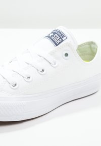 Zapatilla de lona blanca con suela de goma, punta redonda y diseño con cordones. Presenta una etiqueta azul en la lengüeta y sutiles acentos texturizados.